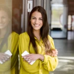 hübsche Businessfrau in gelber Bluse mit weißem Handy an einer Wand angelehnt