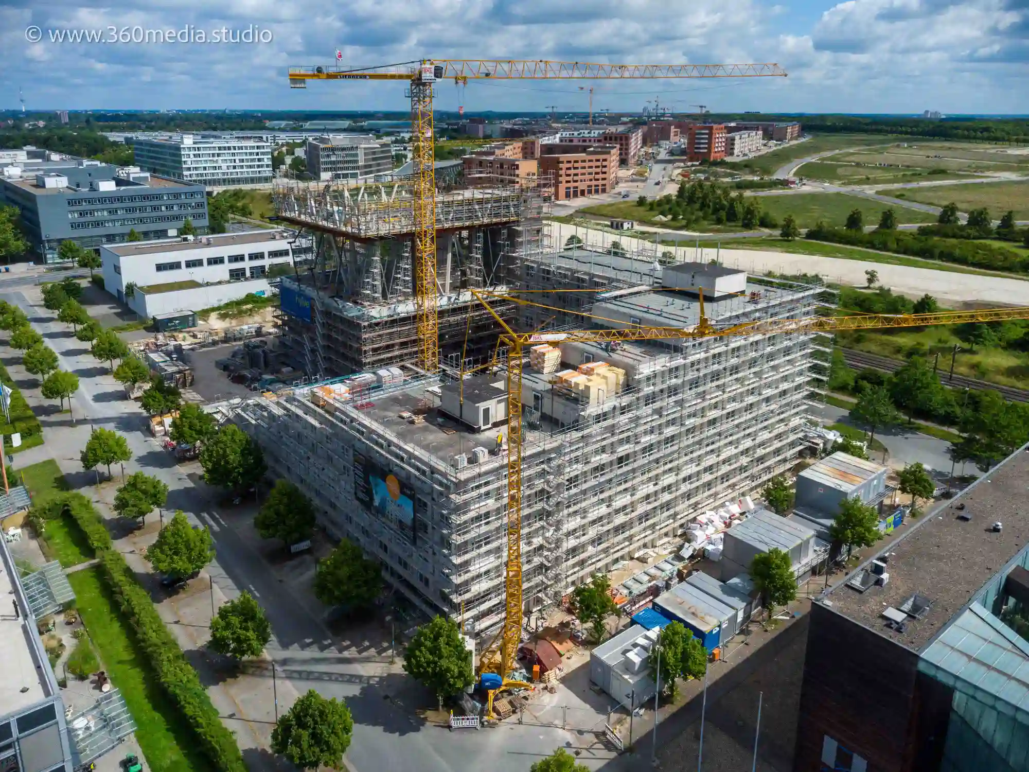 Drohnenaufnahmen aus der Luft zeigen eine Baustelle mit einem hohen gelben Kran, mehreren Gerüsten um ein großes Gebäude im Bau sowie umliegenden Straßen und Gebäuden. Das Gebiet zeigt eine Mischung aus bebautem und unbebautem Land unter klarem Himmel.