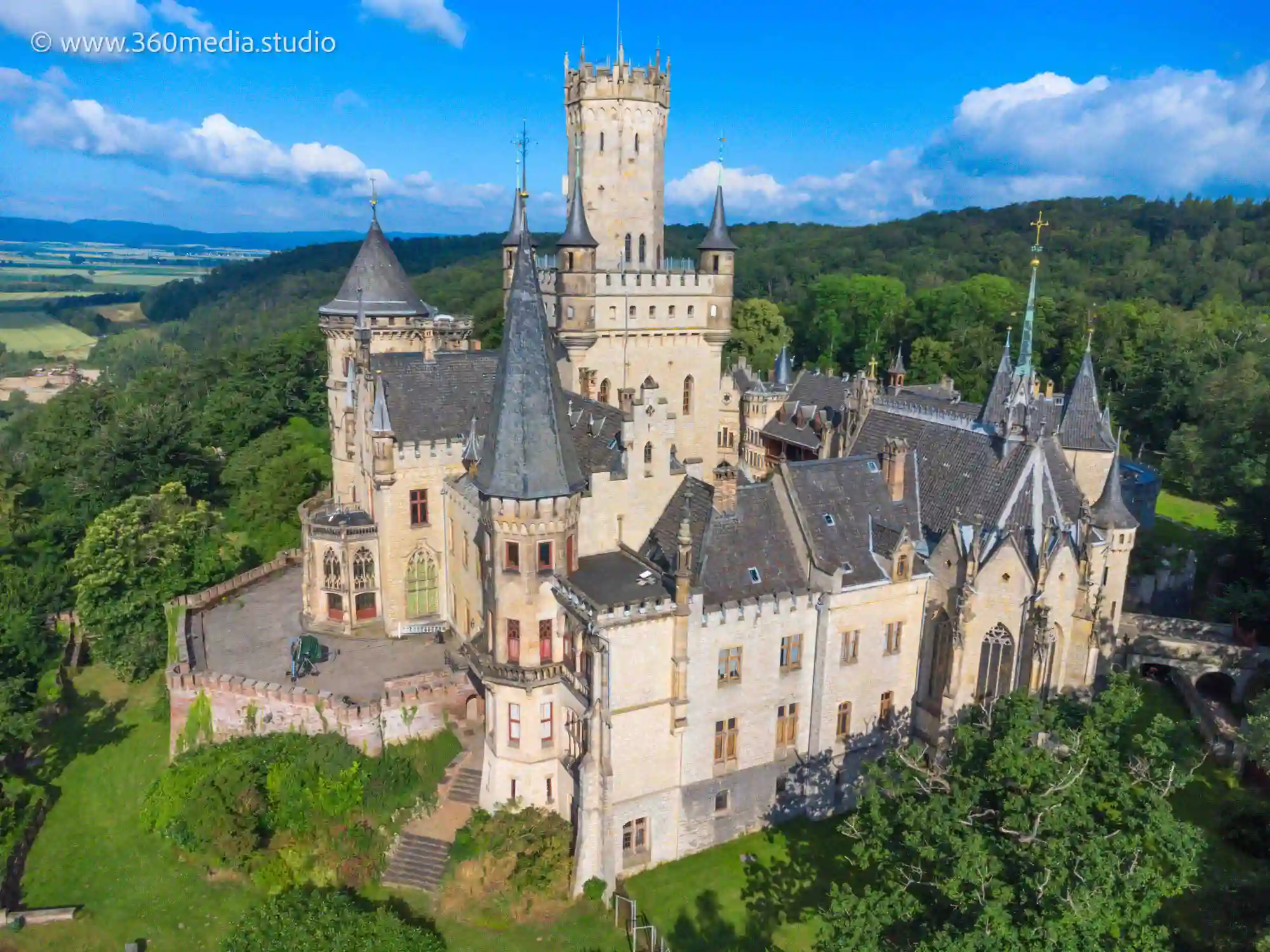 Luftaufnahme des historischen Schlosses Marienburg, umgeben von üppigem Grün in der Region Hannover. Das Schloss verfügt über mehrere Türme, spitze Türme und einen hohen zentralen Turm. Der Himmel ist klar mit ein paar vereinzelten Wolken und die Landschaft in der Ferne umfasst Felder und Hügel.