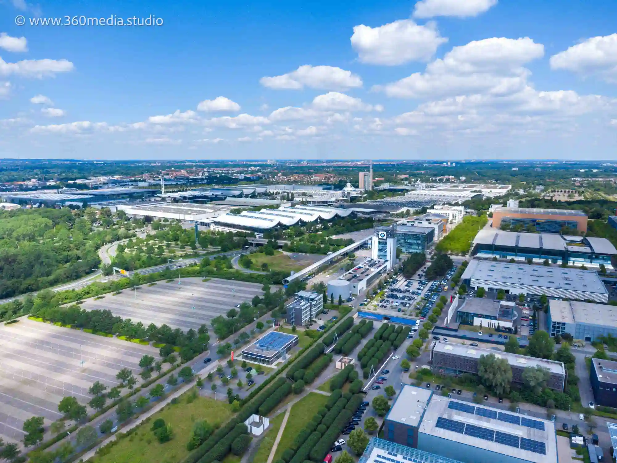 Drohnenaufnahme von einem modernen Vorort in Hannover zeigt eine Mischung aus Wohn- und Gewerbegebieten. Grüne Felder und Bäume sind sichtbar, ebenso Straßen und Parkplätze. Im mittleren linken Bildbereich ist ein Kran nahe einer Baustelle zu sehen, Teil des neuen Stadtteils Kronsrode.