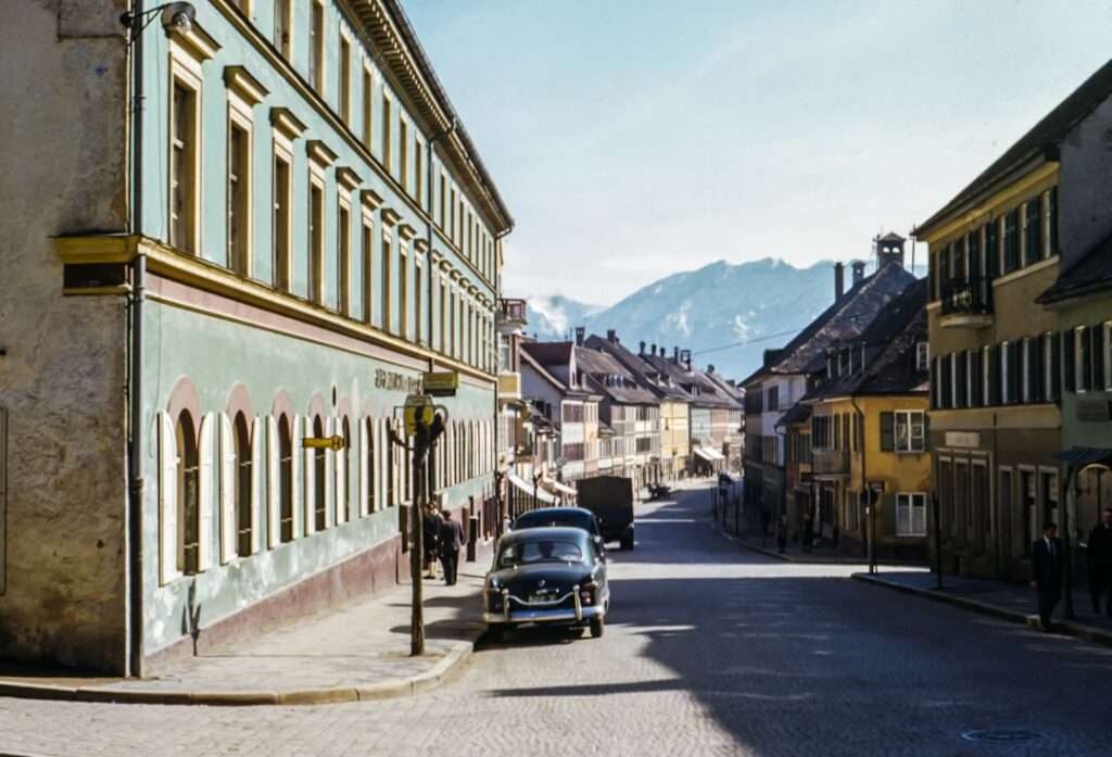 Google Street View Kosten: Was du wissen musst 19 Fotograf Hannover - Eine historische Straßenszene mit einem Oldtimer, der auf einer von historischen Gebäuden gesäumten Kopfsteinpflasterstraße geparkt ist. Im Hintergrund sind Berge unter einem klaren blauen Himmel zu sehen. Schatten deuten auf einen sonnigen Tag hin. Einige Leute sind auf dem Bürgersteig zu sehen. Fotograf in der Nähe.