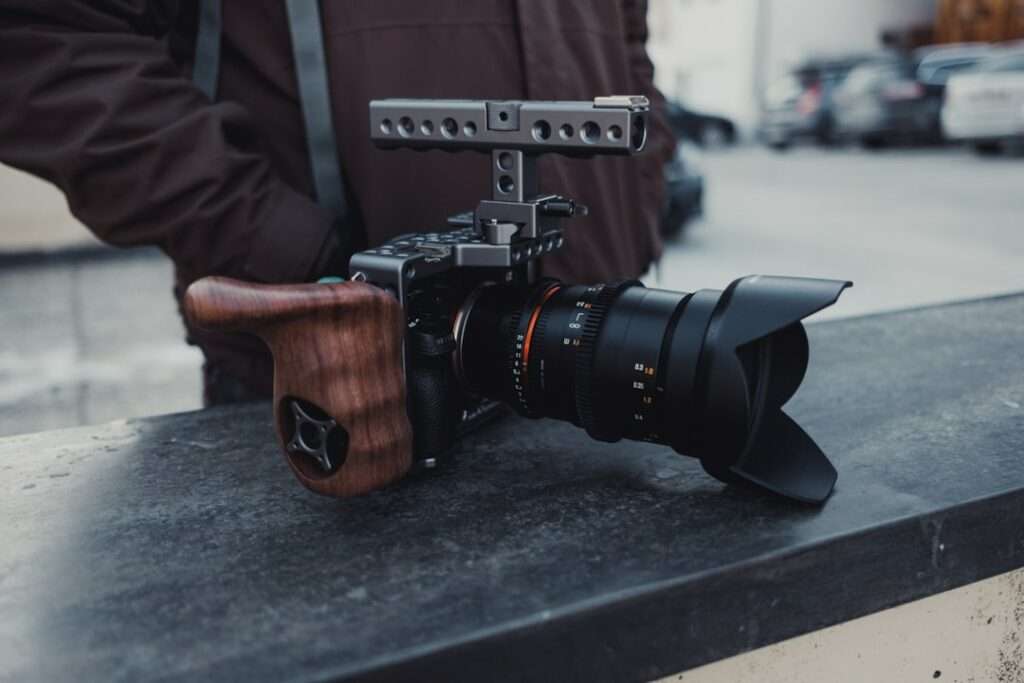 Fotografieren in Hannover: Entdecke die Schönheit mit mir! 16 Fotograf Hannover - Eine Kamera mit Holzgriff und Gegenlichtblende steht auf einer Betonfläche. Dahinter steht eine dunkel gekleidete Person. Im Hintergrund ist ein Parkplatz mit mehreren Fahrzeugen zu sehen. Fotograf in der Nähe.