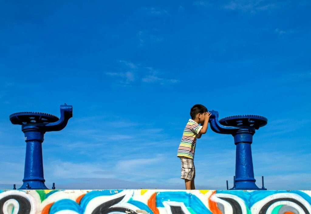 Fotograf Hannover Ein Kind in gestreiftem Hemd und karierten Shorts blickt durch ein blaues Fernrohr, das auf einer bunt bemalten Betonplattform montiert ist. Ein weiteres identisches Teleskop steht neben ihm, als hätte es ein 360-Grad-Fotograf aufgenommen. Im Hintergrund ist der Himmel klar und blau. Fotograf in der Nähe.