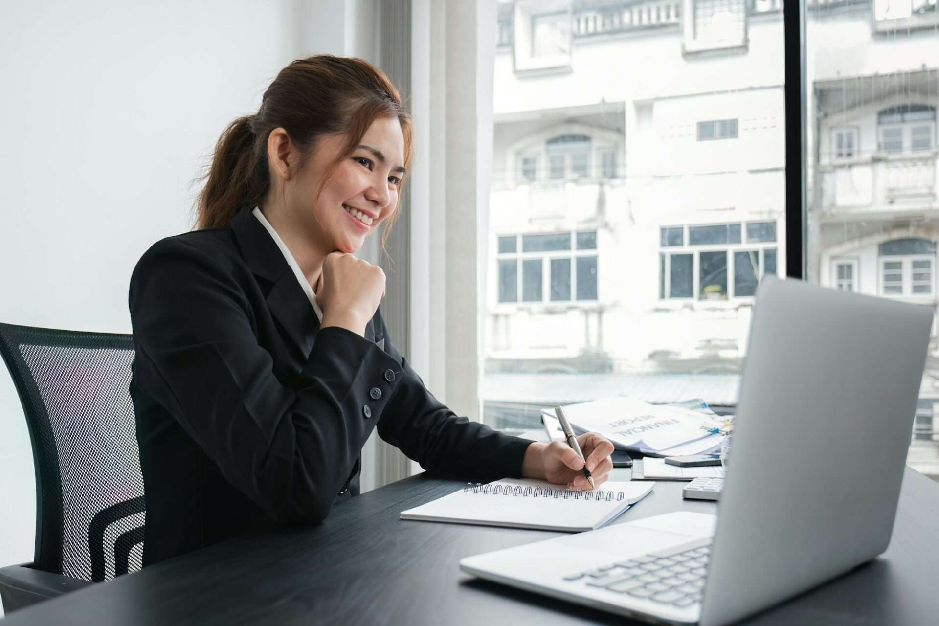 Fotograf Hannover Eine Frau in einem schwarzen Blazer sitzt an einem Schreibtisch vor einem Laptop, lächelt und schreibt in ein Notizbuch. Auf dem Schreibtisch liegen Dokumente, im Hintergrund sieht man große Fenster mit Blick auf die Stadtlandschaft – die Essenz der Unternehmensfotografie. Fotograf in der Nähe.