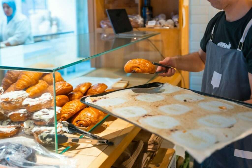 Fotograf Hannover Eine Person in Schürze steht hinter einer Glasvitrine voller Croissants und legt mit einer Zange ein frisch gebackenes Croissant in die Vitrine. In der anderen Hand hält sie ein Tablett mit Backpapier. Der Hintergrund, perfekt eingefangen von einem 360 Grad Fotografen, zeigt eine geschäftige Bäckerei-Umgebung. Fotograf in der Nähe.