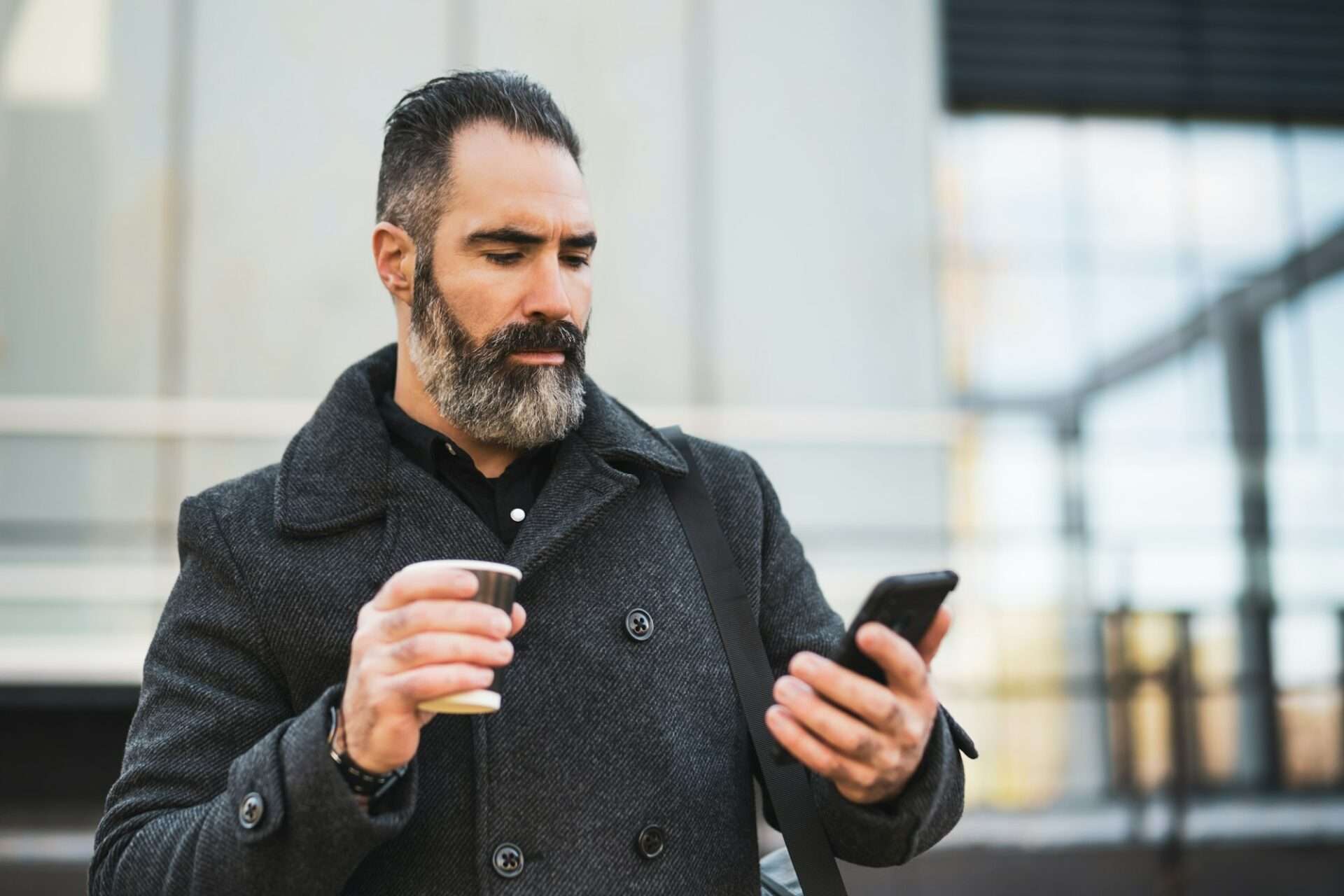 Fotograf Hannover Draußen steht ein bärtiger Mann in einem dunklen Mantel. In der linken Hand hält er einen Pappbecher, in der rechten schaut er auf ein Smartphone. Das Porträt zeigt ihn vor dem Hintergrund von Glasfenstern und einem Teil der Außenfassade des Gebäudes. Fotograf in der Nähe.