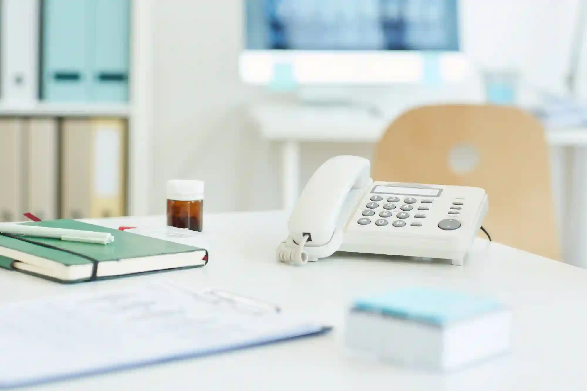 Fotograf Hannover Ein weißer Bürotisch mit einem Telefon, einem grünen Notizbuch mit einem Bleistift darauf, einer kleinen braunen Medikamentenflasche und einem Stapel Papier. Im Hintergrund ein Holzstuhl und ein Computer mit unscharfem Bildschirm, der typische Praxisfotos einfängt. Fotograf in der Nähe.