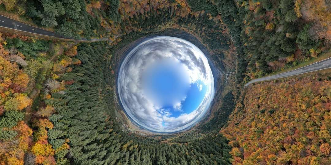 Fotograf Hannover Ein Luftbild eines dichten Waldes mit einem sphärischen Verzerrungseffekt in der Mitte, der den Himmel als runden Brennpunkt erscheinen lässt. Zwei Straßen mit herbstlich gefärbten Bäumen auf beiden Seiten säumen den Wald. Das Bild wirkt surreal, tunnelartig, perfekt für eine Online-Besichtigung. Fotograf in der Nähe.