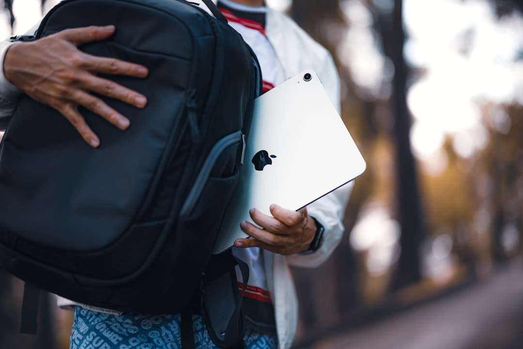 Fotograf Hannover Eine Person in einer weißen Jacke steckt ein Apple iPad in einen schwarzen Rucksack und fängt damit eine Szene ein, die sich ideal für Produktfotografie eignet. Im Hintergrund ist ein von Bäumen gesäumter Weg zu sehen, der jedoch unscharf ist. Fotograf in der Nähe.