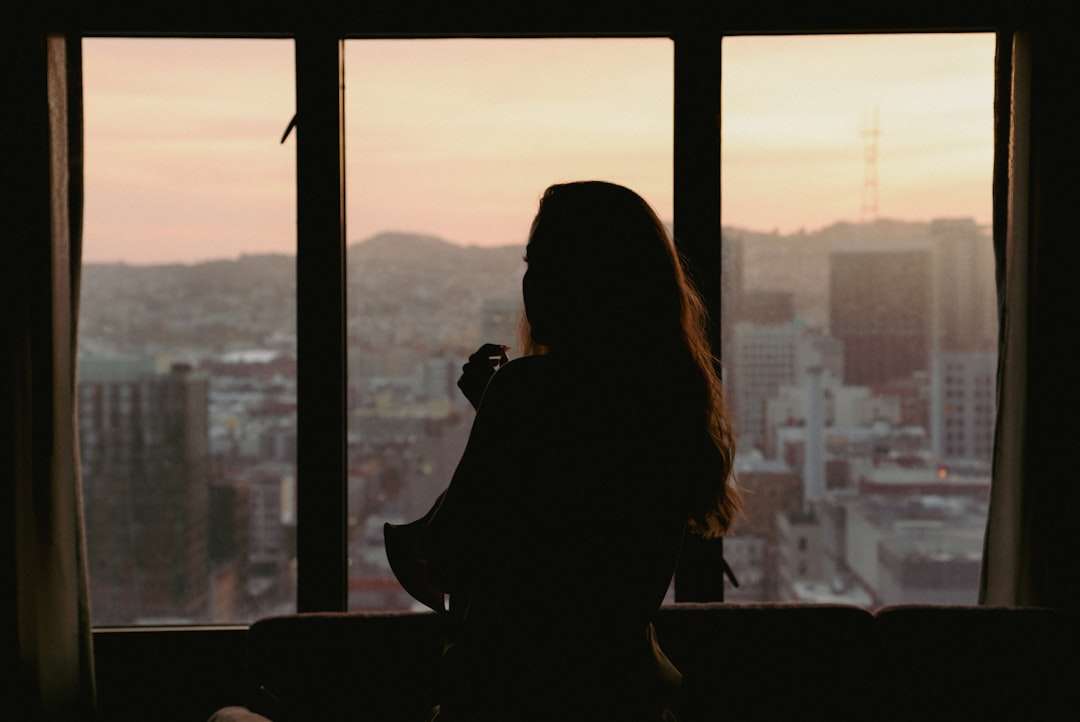 Fotograf Hannover Eine Person mit langen Haaren ist als Silhouette vor einem großen Fenster in ihrem Hotelzimmer zu sehen und blickt bei Sonnenuntergang auf eine Stadtlandschaft. Der Raum ist schwach beleuchtet und die Außenszene zeigt Gebäude und entfernte Hügel unter einem Himmel mit Farbverlauf, was einen Moment perfekter Fotografie einfängt. Fotograf in der Nähe.