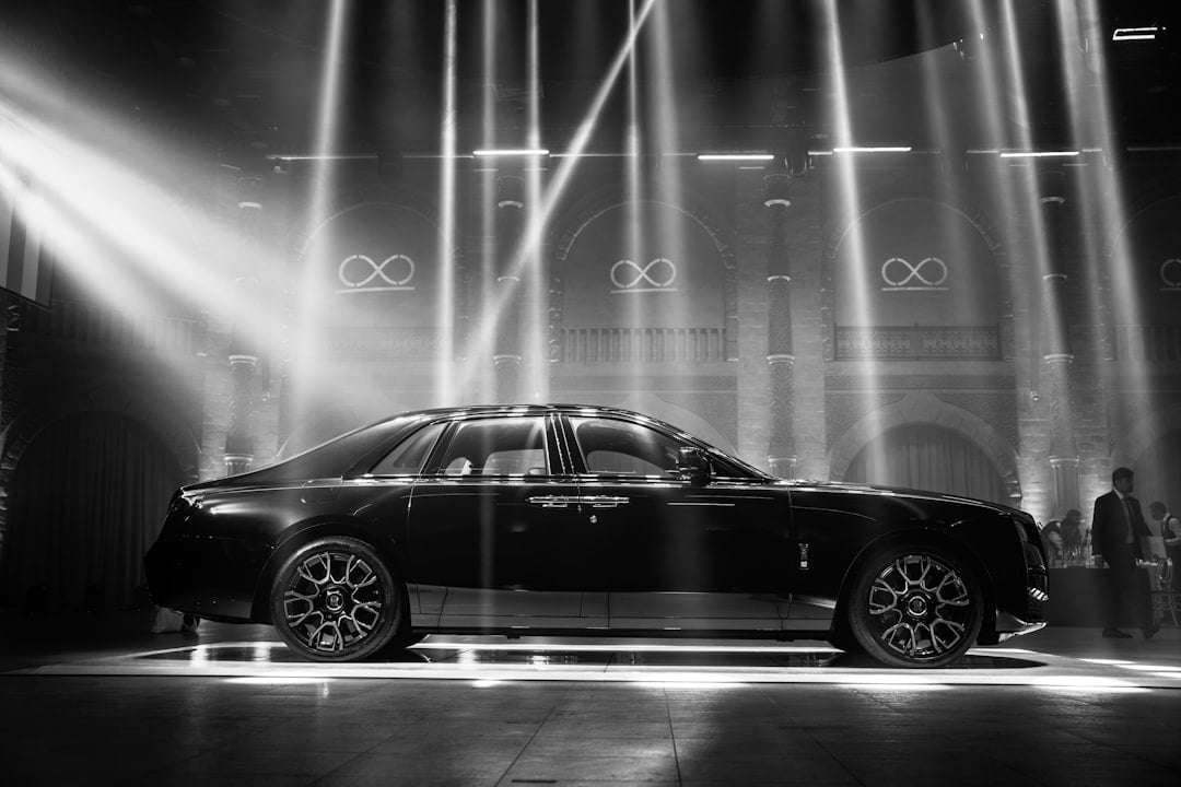 Fotograf Hannover Eine schwarze Luxuslimousine wird in einem Innenbereich unter mehreren hellen Scheinwerfern ausgestellt. Die Szene ist dramatisch, mit Lichtstrahlen, die durch die leicht neblige Luft schneiden und das schlanke und elegante Erscheinungsbild des Fahrzeugs betonen. Der Hintergrund zeigt gewölbte Fenster und beleuchtete Symbole, die an die sorgfältige Einrichtung eines Autofotografen erinnern. Fotograf in der Nähe.