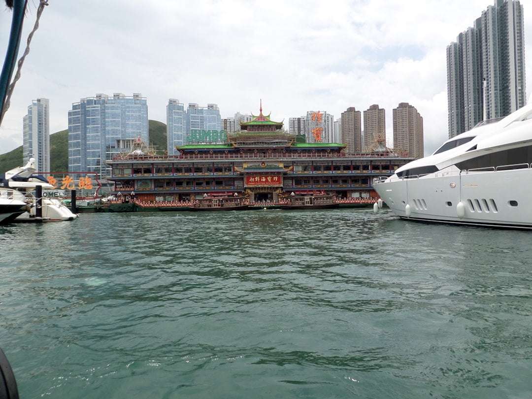 Fotograf Hannover Ein großes schwimmendes Restaurant mit traditionell chinesischer Architektur liegt in einem Hafen vor Anker, flankiert von zwei Luxusyachten, alles vor einer Kulisse aus Hochhäusern. Der Himmel ist teilweise bewölkt, perfekt für diejenigen, die eine „virtuelle Tour erstellen lassen“ und die atemberaubende Landschaft online erleben möchten. Fotograf in der Nähe.