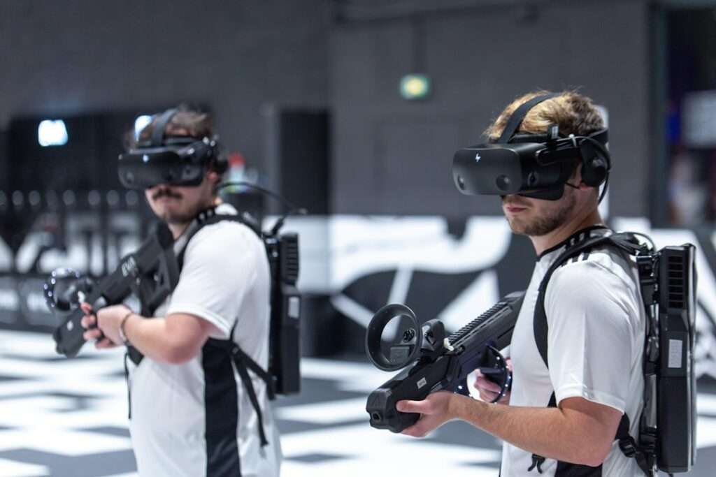 Entdecke die Welt mit einer 360-Grad-Tour 4 Fotograf Hannover Zwei Personen sind in ein Virtual-Reality-Spiel vertieft. Sie tragen VR-Headsets und halten VR-Controller in Pistolenform in der Hand. In weißen Hemden und schwarzen Gamer-Westen gekleidet navigieren sie durch eine monochrome Indoor-Gaming-Umgebung, die von einem 360-Grad-Fotografen aufgenommen wurde. Fotograf in der Nähe.