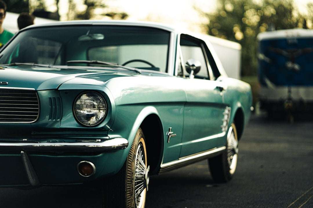 Fotograf Hannover Ein türkisfarbener Ford Mustang steht auf einer gepflasterten Fläche. Das Auto hat ein klassisches Design mit einem Chromgrill und runden Scheinwerfern. Im Hintergrund sind Bäume und ein Gebäude zu sehen. Das Foto wurde schräg von vorne links vom Fahrzeug aufgenommen. Fotograf in der Nähe.