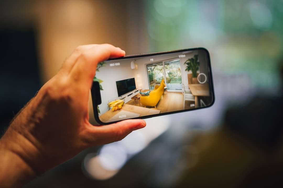 Fotograf Hannover Eine Hand hält ein Smartphone und macht ein Foto von einem modernen Wohnzimmer. Der Raum verfügt über eine leuchtend gelbe Couch, einen an der Wand montierten Fernseher und große Fenster, die natürliches Licht hereinlassen. Zimmerpflanzen und ein Couchtisch sind ebenfalls zu sehen, perfekt für jemanden, der einen virtuellen Rundgang erstellen möchte. Fotograf in der Nähe.