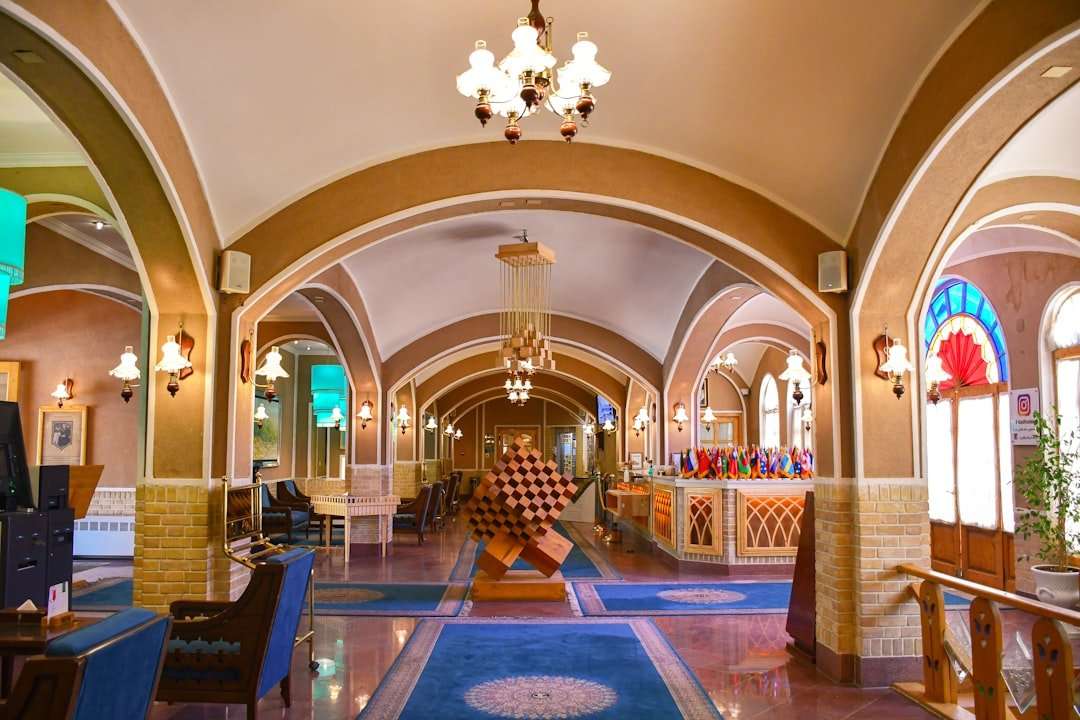 Fotograf Hannover Eine Hotellobby mit gewölbten Decken und Kronleuchtern, in deren Mitte sich eine geometrische Holzskulptur befindet. Im Hintergrund sind an einem Check-in-Schalter verschiedene Flaggen zu sehen. An den Wänden befinden sich Sitzbereiche und dekorative Lampen. Fotograf in der Nähe.