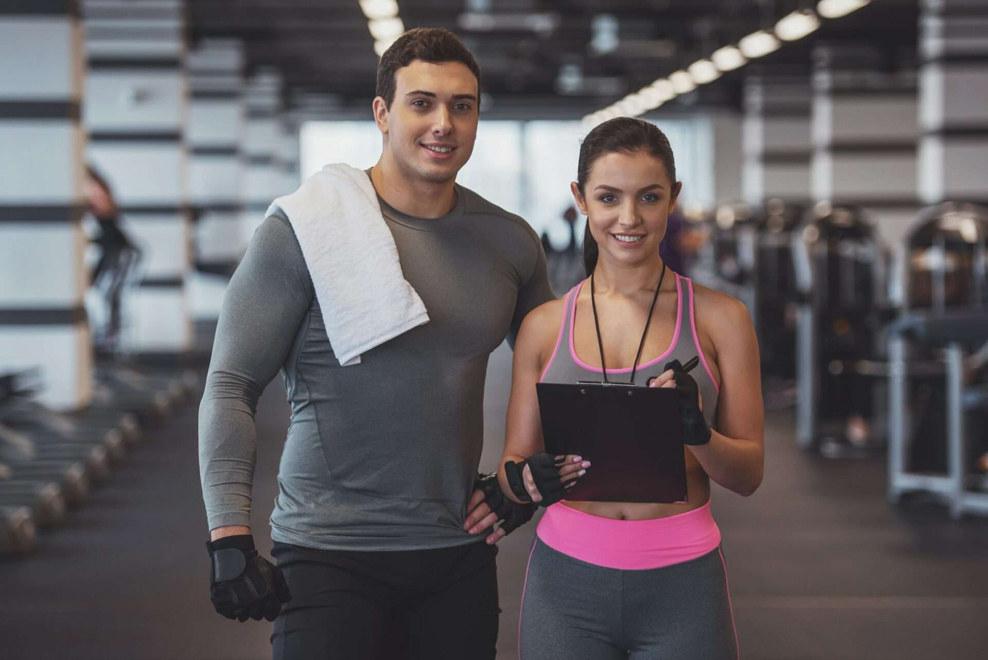 Fotograf Hannover Zwei Personen in Sportkleidung stehen in einem Fitnessstudio. Der Mann links hat ein Handtuch über der Schulter und die Frau rechts hält ein Klemmbrett. Beide lächeln. Im Hintergrund sind Trainingsgeräte zu sehen. Fotograf in der Nähe.