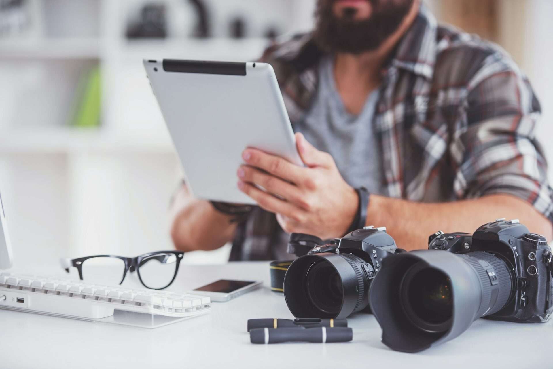 Fotograf Hannover Ein 360-Grad-Fotograf sitzt an einem Schreibtisch, der mit verschiedenen Gegenständen wie einer Tastatur, einer Brille, zwei DSLR-Kameras und einem Objektivdeckel ausgestattet ist, und verwendet ein Tablet. Die Person trägt ein kariertes Hemd und eine schicke Uhr. Fotograf in der Nähe.