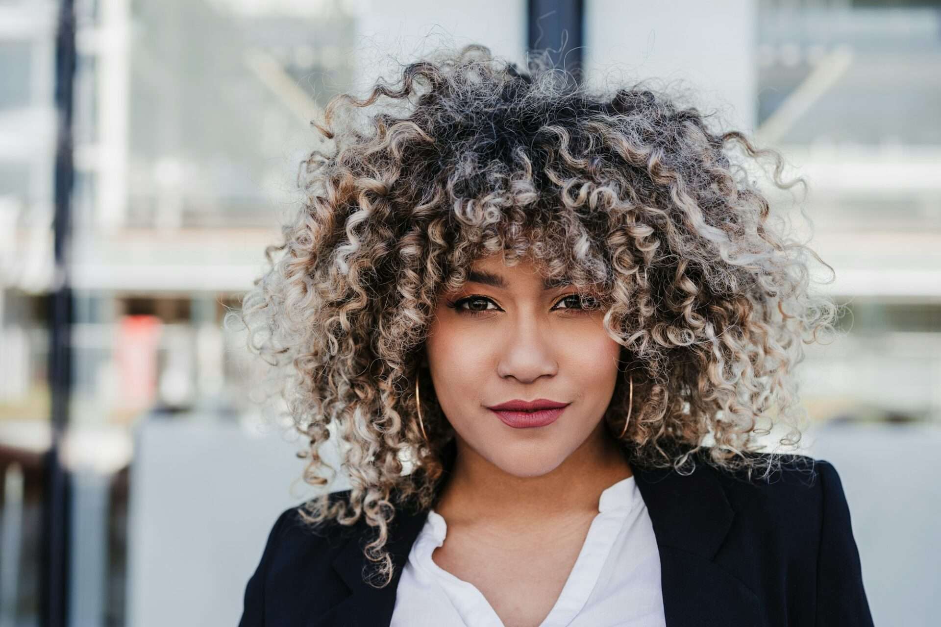 Fotograf Hannover Eine Frau mit lockigem Haar steht im Freien und blickt in die Kamera, um ein professionelles Geschäftsporträt aufzunehmen. Sie trägt einen schwarzen Blazer über einem weißen Oberteil und hat einen neutralen Gesichtsausdruck. Der Hintergrund ist verschwommen und zeigt ein Gebäude mit großen Fenstern. Fotograf in der Nähe.