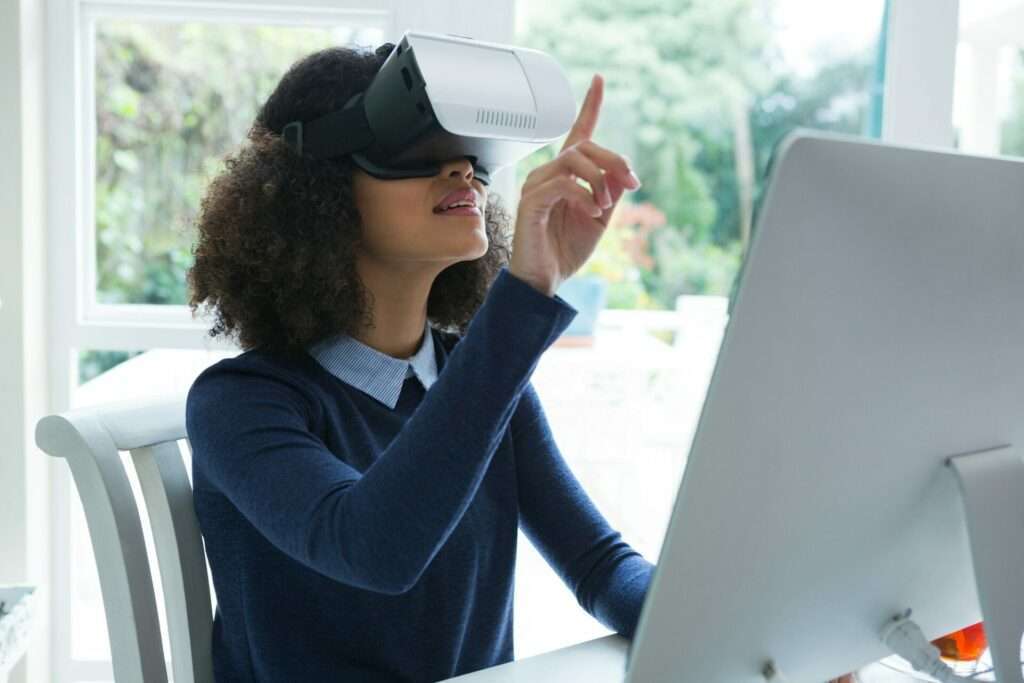 Entdecke dein neues Zuhause mit virtueller Besichtigung 28 Fotograf Hannover Eine Person sitzt an einem Schreibtisch und benutzt einen Desktop-Computer, während sie ein Virtual Reality (VR)-Headset trägt, und nimmt an einer virtuellen Besichtigung teil. Ihre Hand ist erhoben und zeigt nach vorne, als würde sie mit der virtuellen Umgebung interagieren. Im Hintergrund ist ein großes Fenster mit Grünflächen zu sehen. Fotograf in der Nähe.