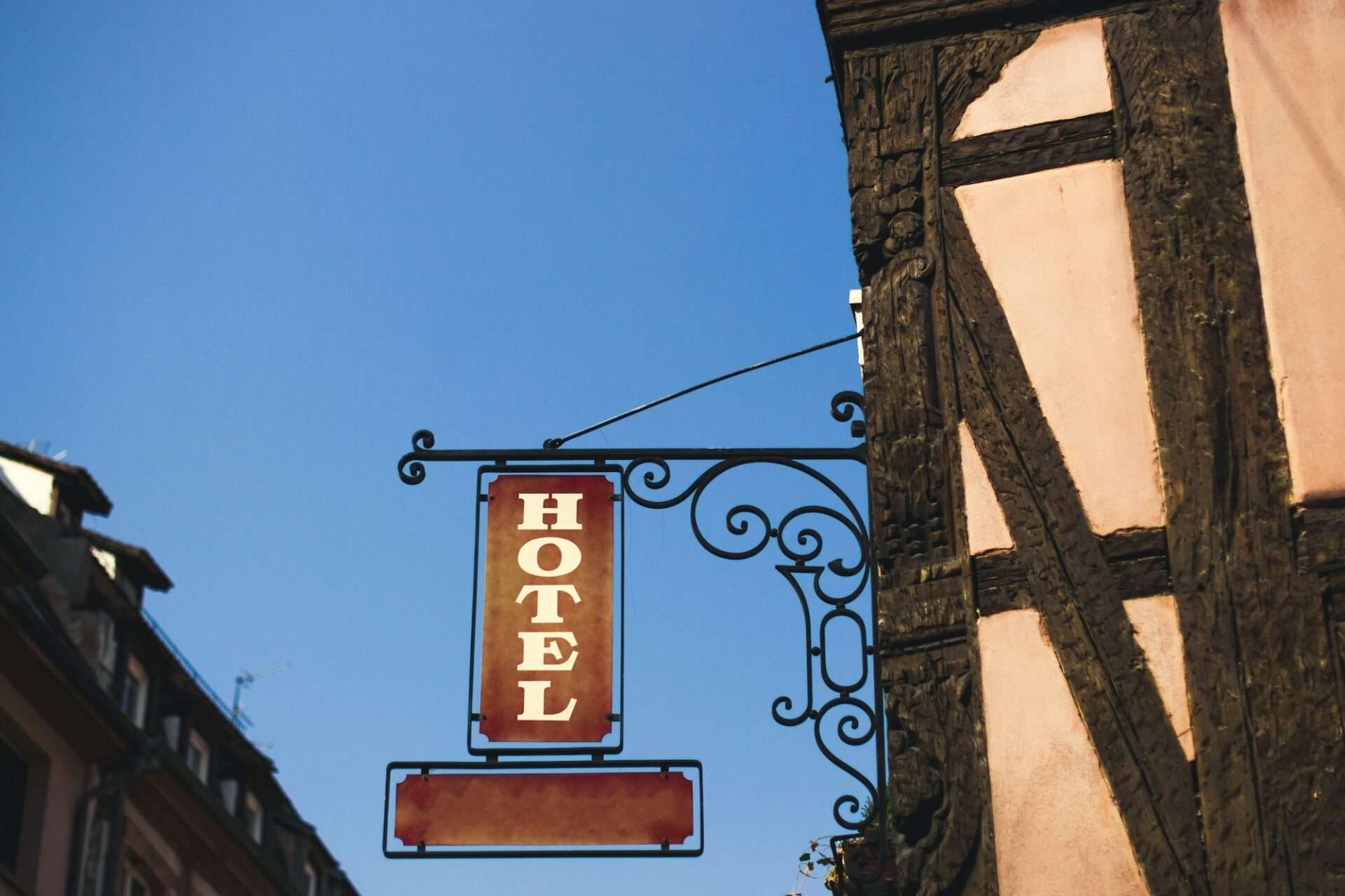 Fotograf Hannover Ein Hotelschild im Vintage-Look hängt an einer verzierten Halterung an einem Fachwerkhaus – perfekt für jeden Hotelfotografen. Auf dem Schild steht in weißen Buchstaben „HOTEL“ auf rotem Hintergrund und der Himmel ist klar und blau. Fotograf in der Nähe.