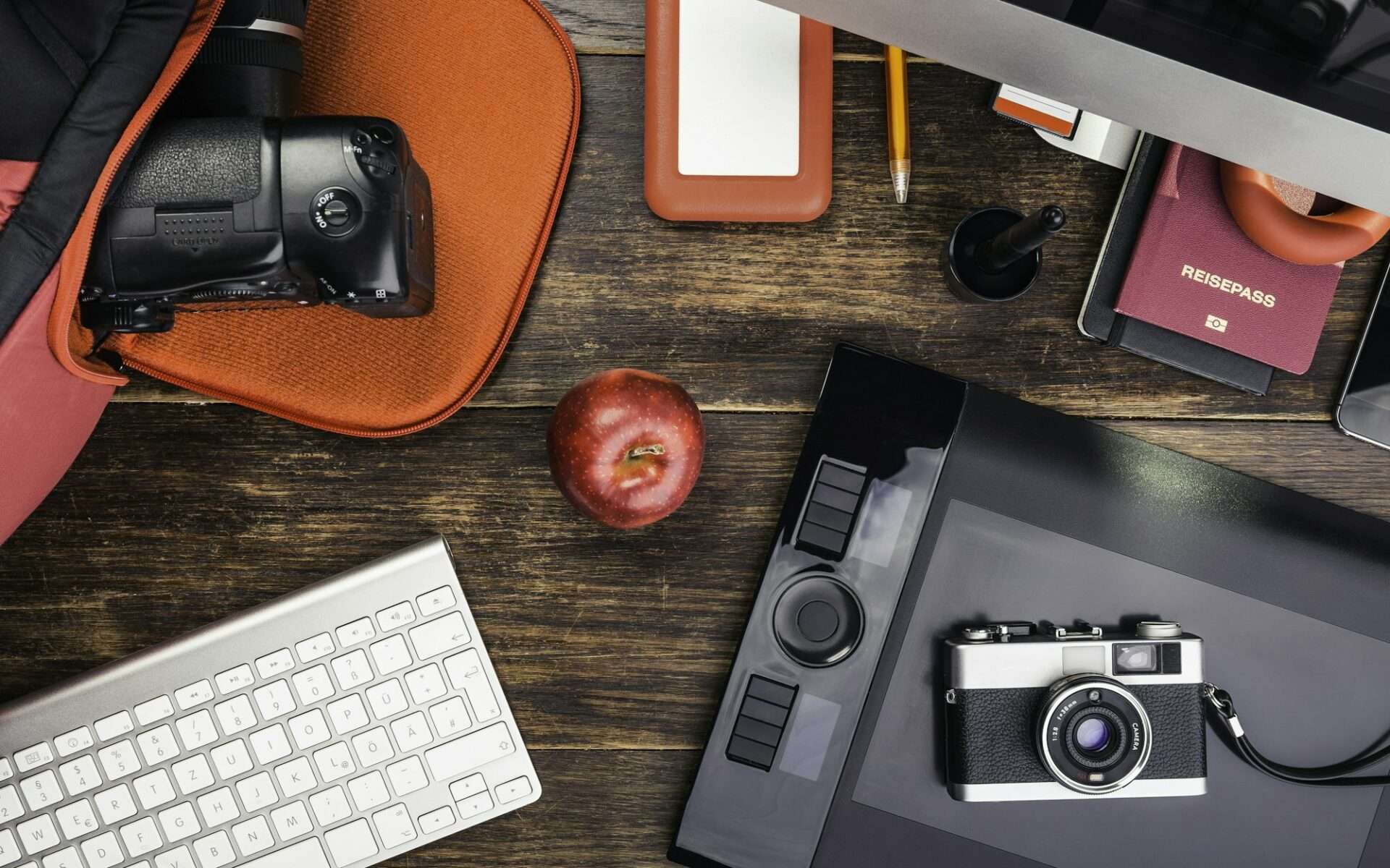 Fotograf Hannover Auf einem Holzschreibtisch stehen eine Kamera, ein digitales Tablet, eine Tastatur und eine externe Festplatte für die nahtlose Bildbearbeitung. Daneben liegen ein Apfel, ein Stift, ein Bleistift, ein roter Reisepass, ein Computermonitor und eine orange Laptophülle mit einer weiteren teilweise sichtbaren Kamera. Fotograf in der Nähe.