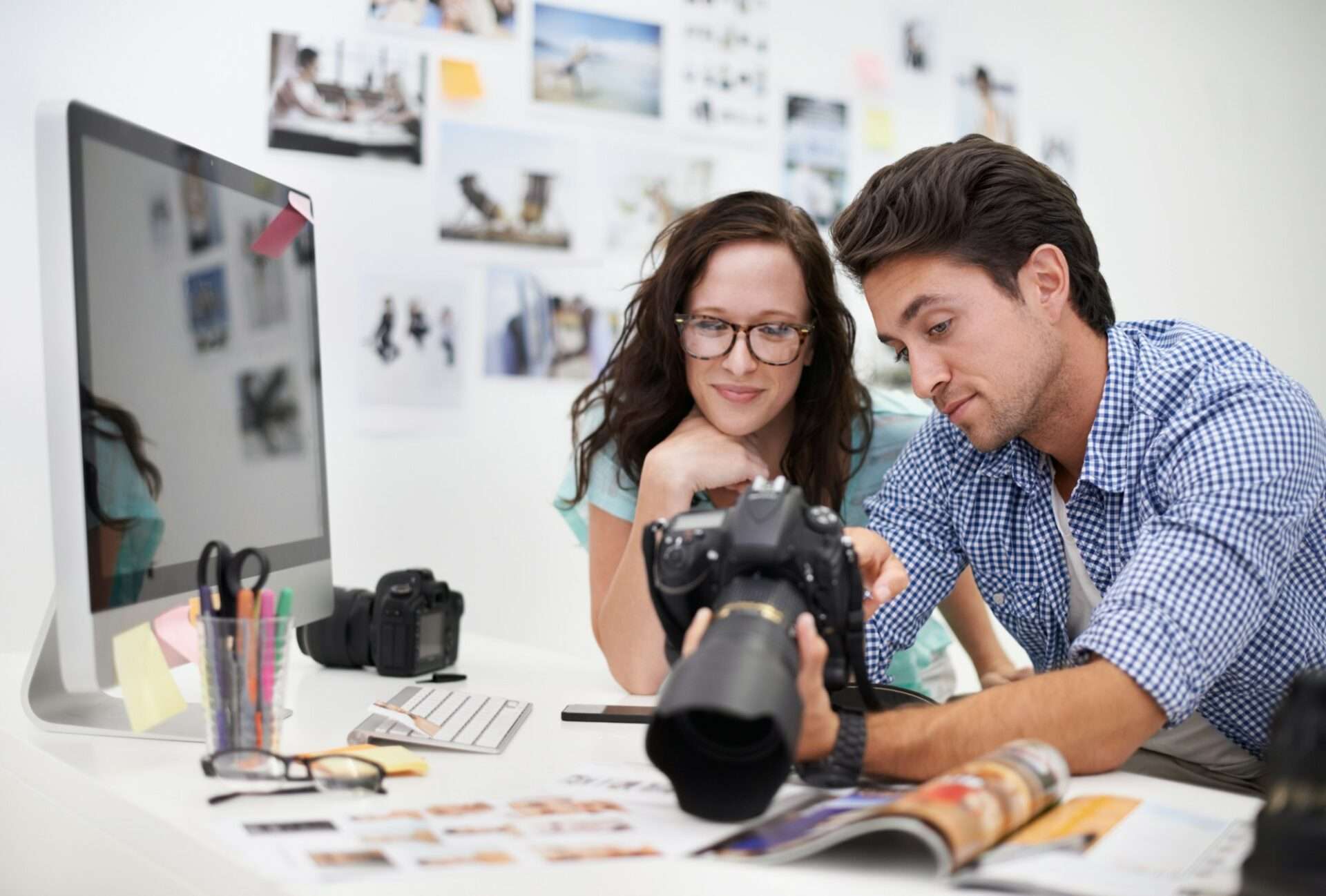 Fotograf Hannover Ein Mann und eine Frau untersuchen eine Digitalkamera in einem Büro, wo Geschäftsfotos die Wand schmücken. Auf dem Schreibtisch stehen ein Computer, ein Kameraobjektiv, Stifte und Fotos. Sie sind leger gekleidet und konzentrieren sich auf ihr gemeinsames Interesse an der Fotografie. Fotograf in der Nähe.