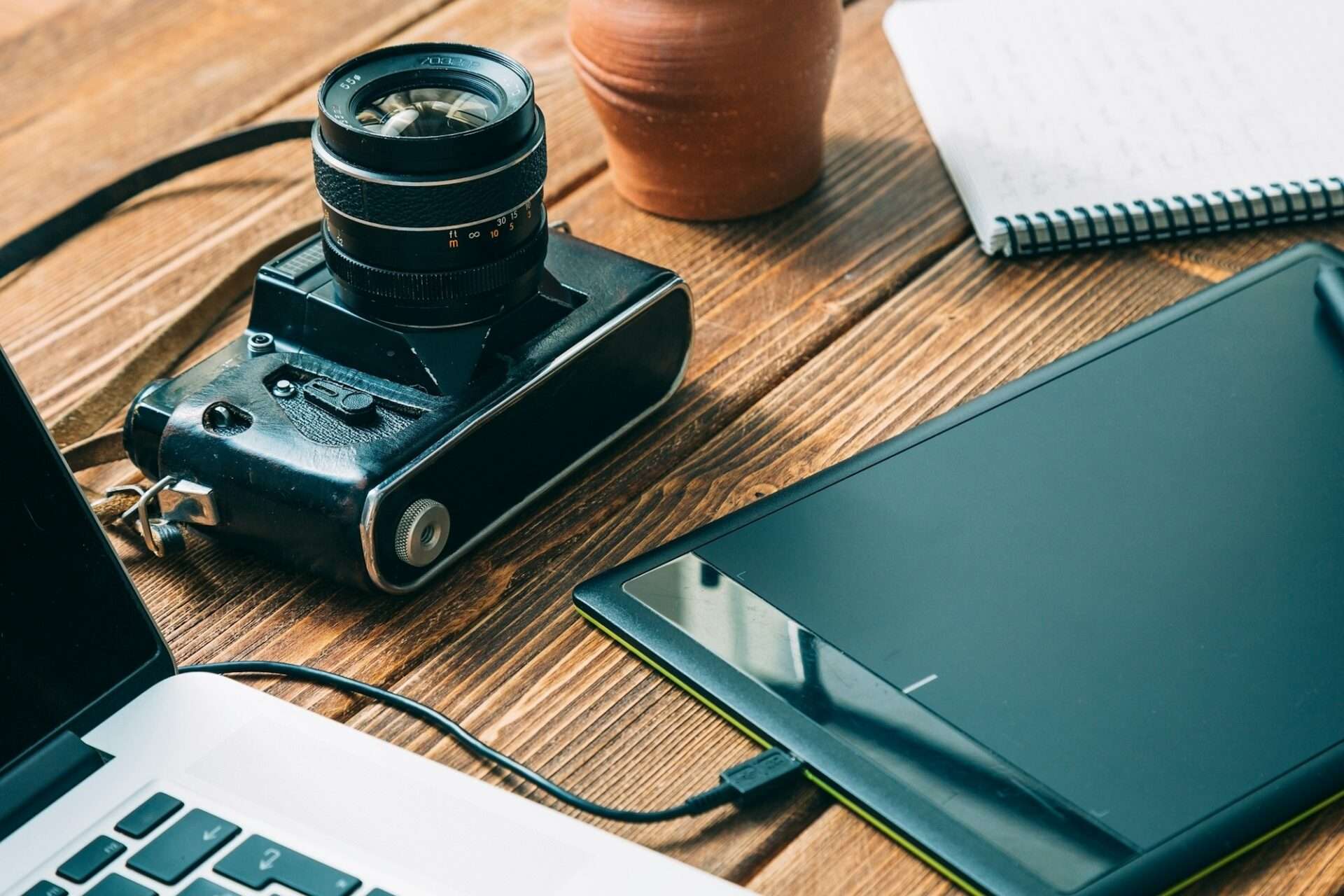 Fotograf Hannover Eine Vintage-Kamera, ein digitales Zeichentablett und ein geöffneter Laptop stehen auf einem Holztisch. Im Hintergrund ist neben einem kleinen braunen Topf ein Notizbuch mit handschriftlichen Notizen teilweise sichtbar – perfekt für einen 360-Grad-Fotografen, der kreative Inspiration einfängt. Fotograf in der Nähe.