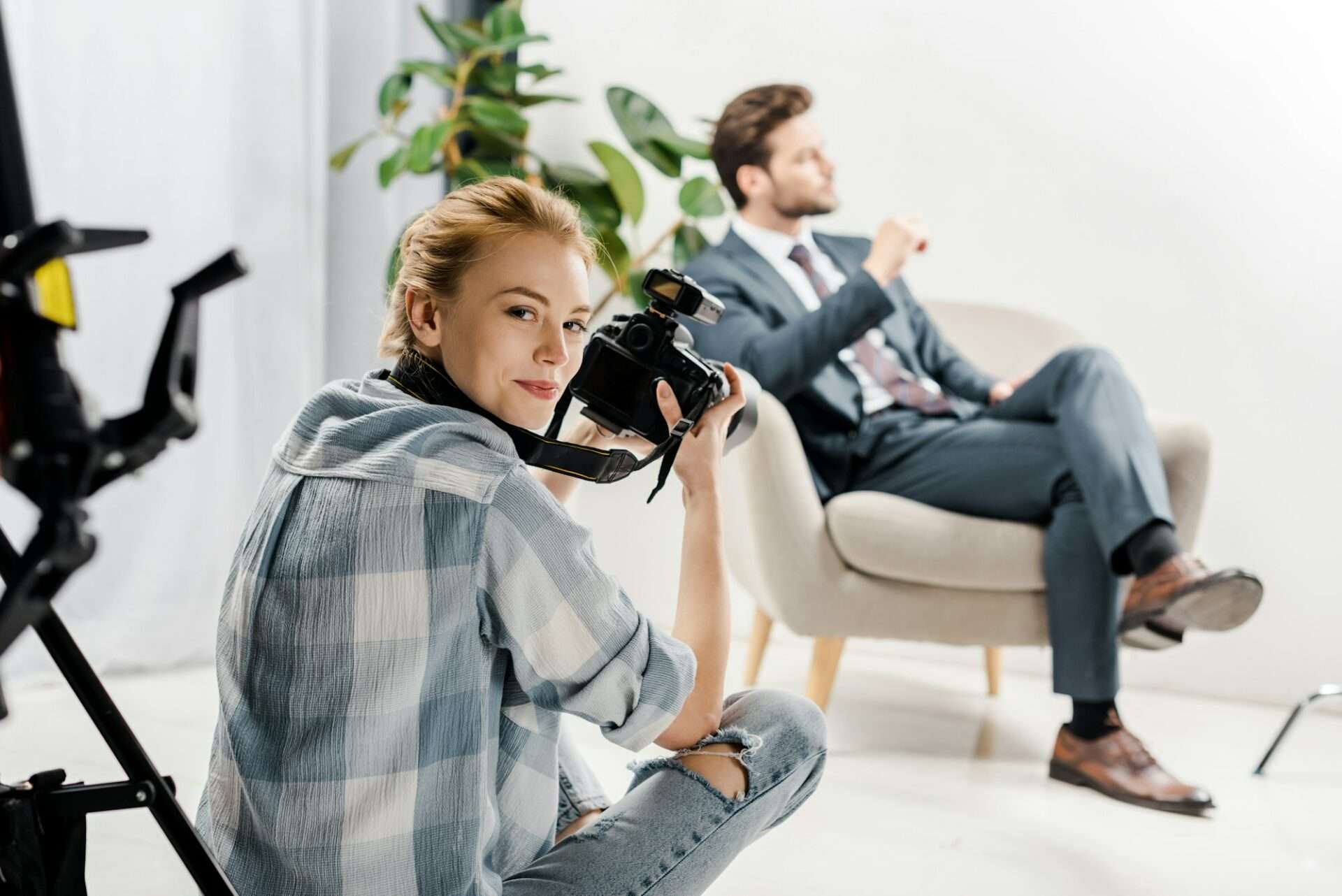 Fotograf Hannover Eine Frau mit einer Kamera sitzt auf dem Boden und lächelt in die Linse, als wäre sie bereit für ihr nächstes 360-Grad-Fotoshooting. Im Hintergrund sitzt ein Mann im Anzug auf einem Sessel, leicht verschwommen inmitten grüner Pflanzen und teilweise sichtbarer Studiobeleuchtungsausrüstung. Fotograf in der Nähe.