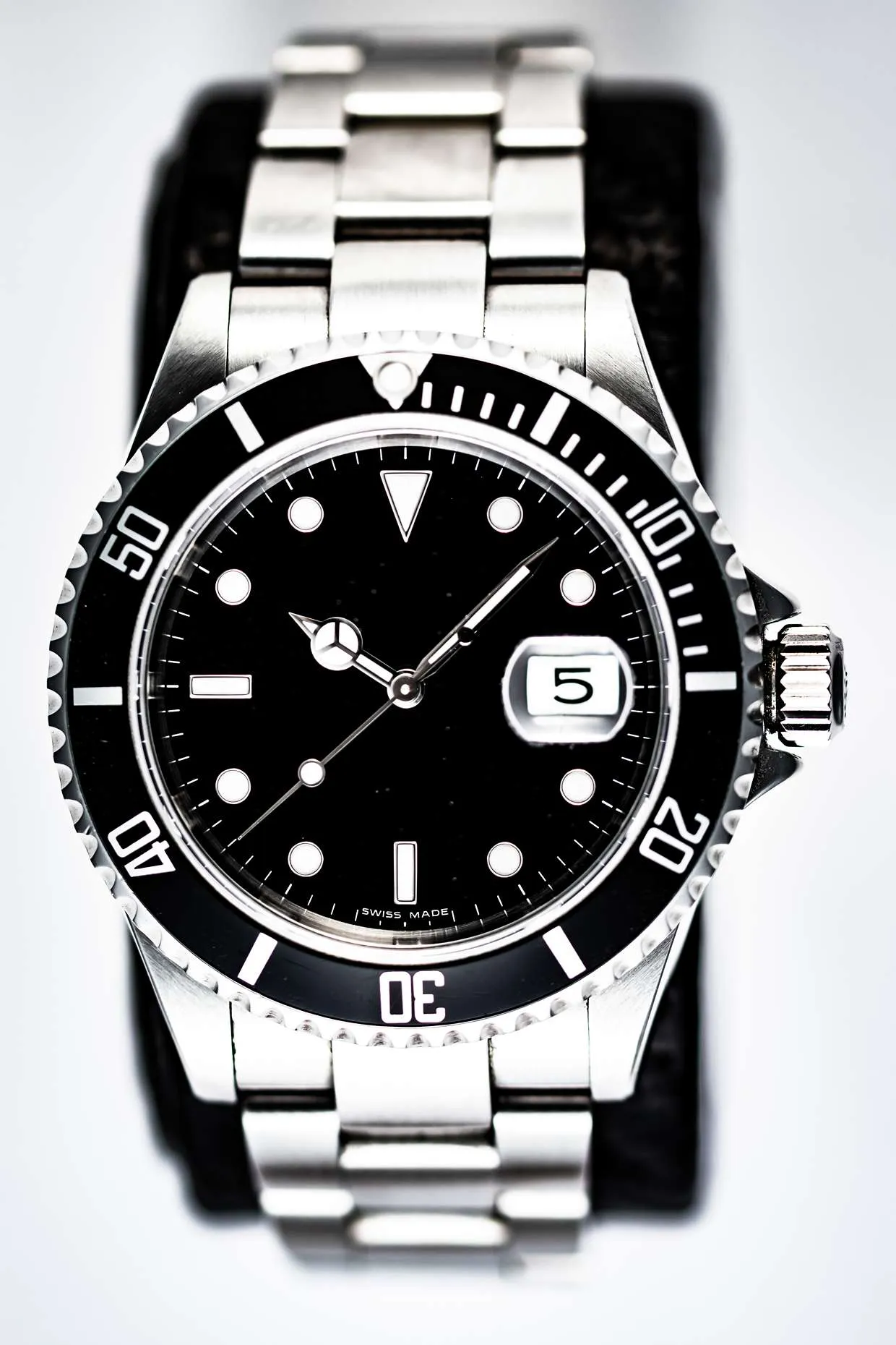 Produktfotos 8 Fotograf Hannover - Eine Nahaufnahme von Werbefotograf Hannover einer Edelstahlarmbanduhr mit schwarzem Zifferblatt, weißen Stundenmarkierungen, drehbarer Lünette und Metallarmband. Die Datumsanzeige zeigt die Zahl 5. Fotograf in der Nähe.